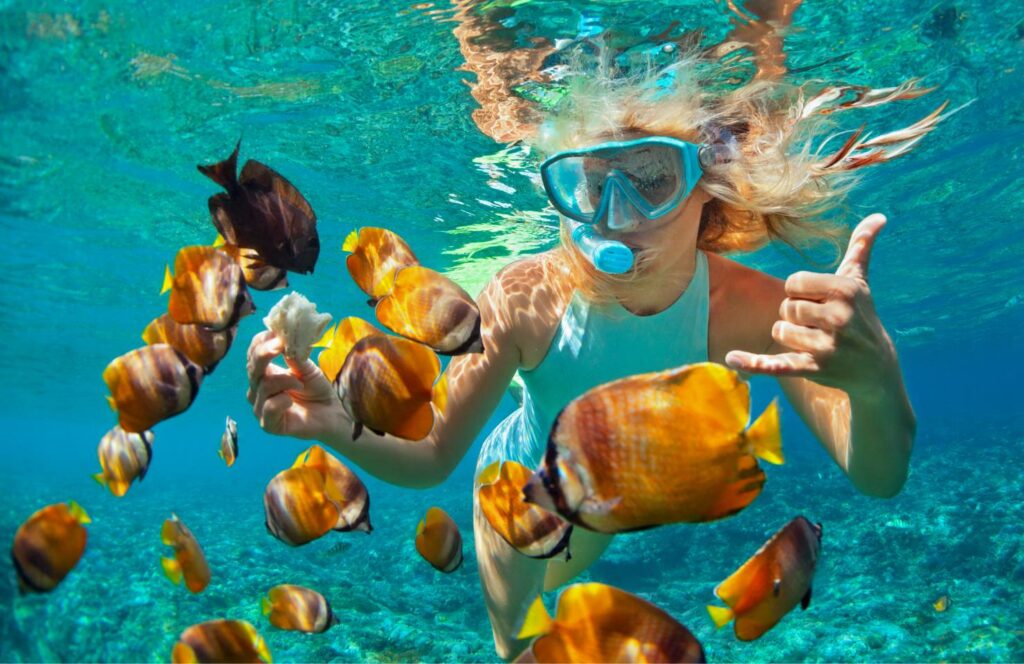 Une femme fait de la plongée avec tuba entourée de poissons orange dans une eau turquoise