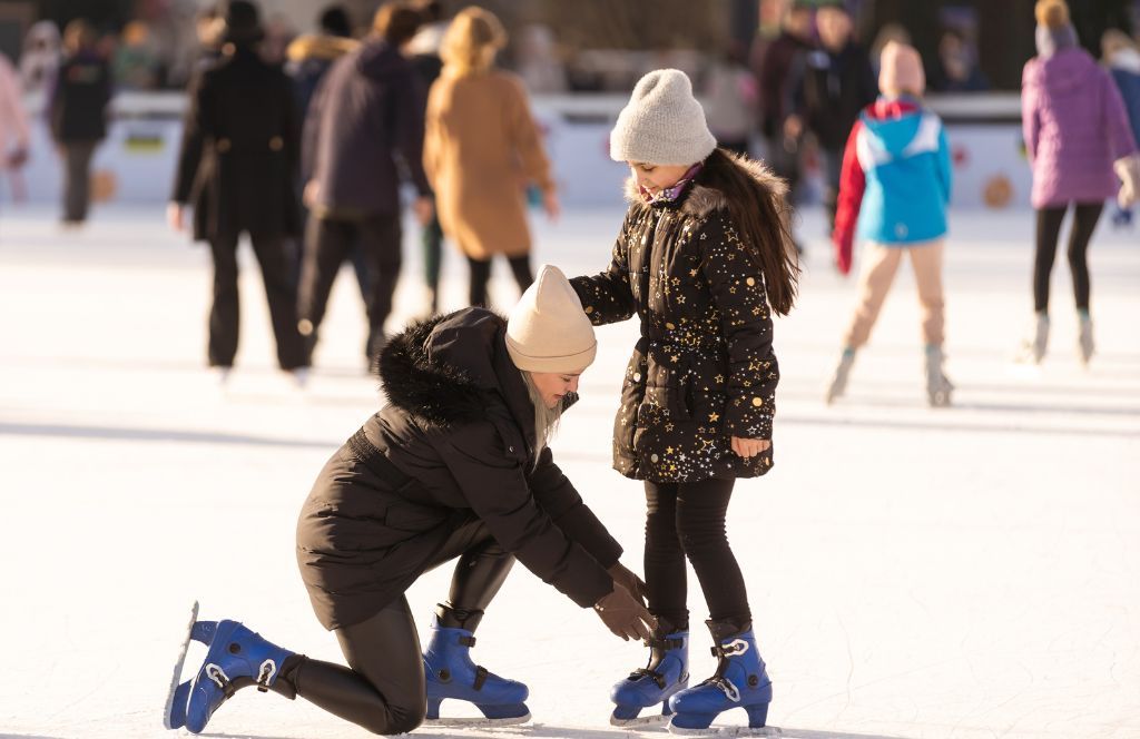 eine Mutter hilft ihrer Tochter beim Einstellen der Schlittschuhe, während sie auf einer Eisbahn in England Schlittschuh laufen - eines der besten britischen Weihnachtserlebnisse